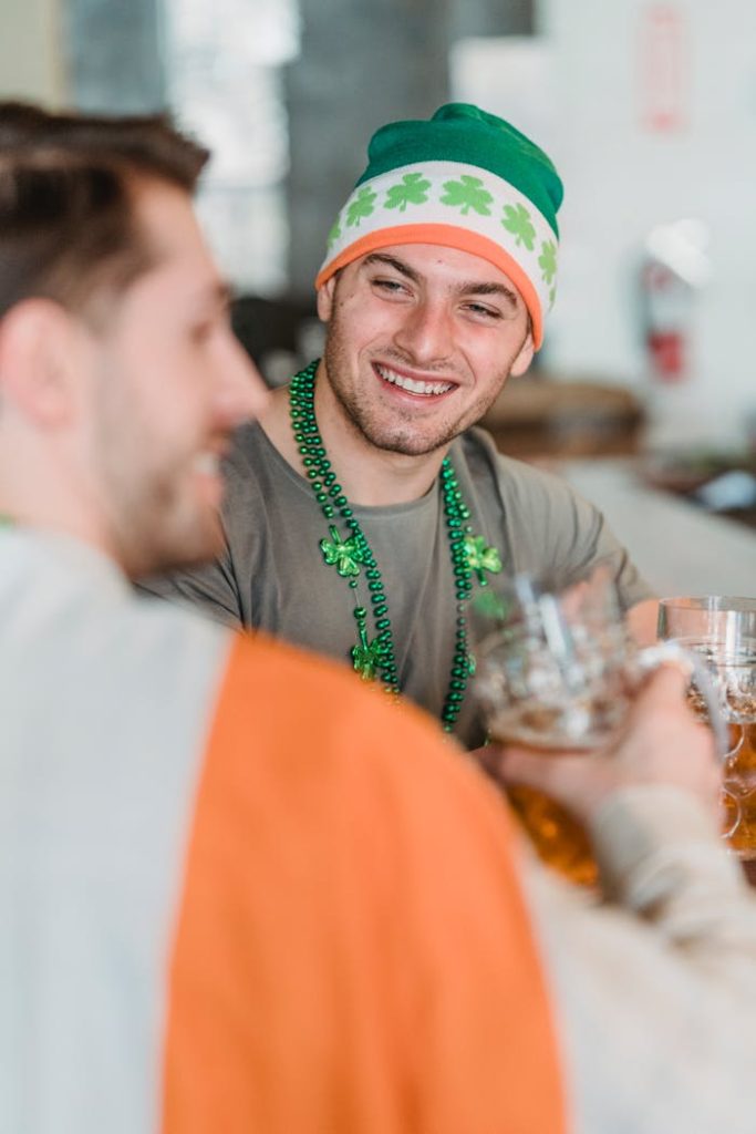 a-man-wearing-beanie-while-smiling-7084029 Two men enjoying beers, celebrating St Patricks Day in festive attire indoors.